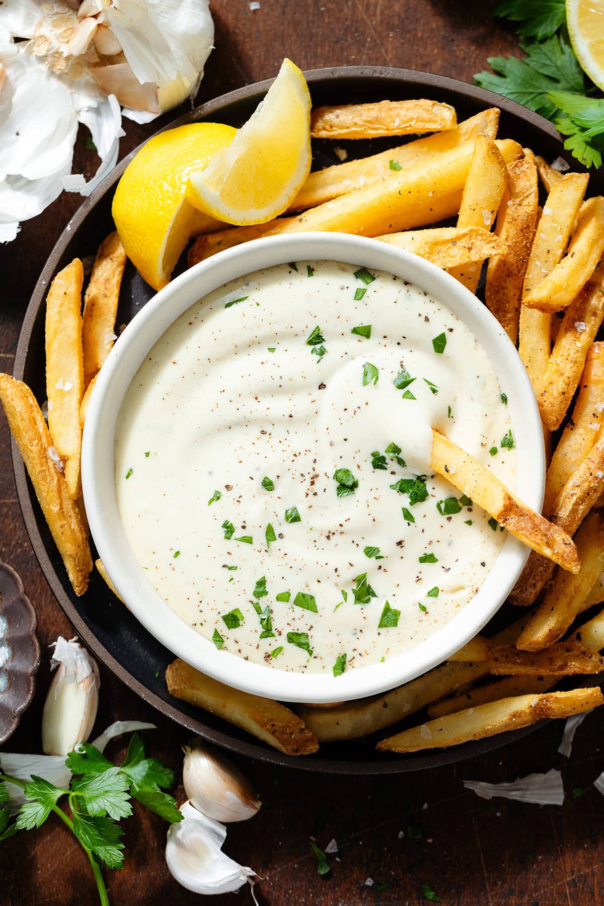 Creamy white dip in a small white bowl garnished with fresh parsley on a black plate with french fries with one fry in the dip.