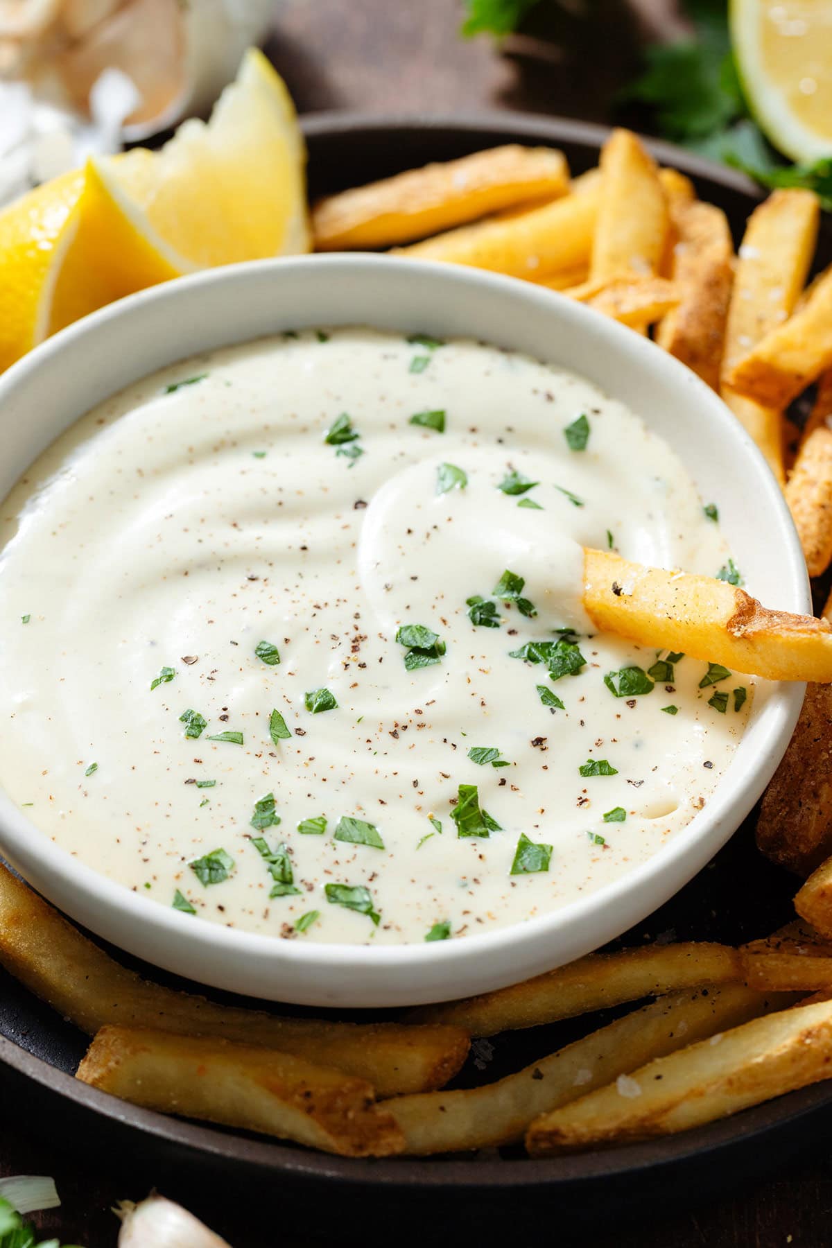 Creamy white dip in a small white bowl garnished with fresh parsley on a black plate with french fries with one fry in the dip.