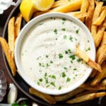 Creamy white dip in a small white bowl garnished with fresh parsley on a black plate with french fries with one fry in the dip.