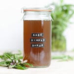 A light brown sage syrup in a tall glass jar with a glass lid with fresh sage around the jar.