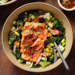 A bowl of kale salad with edamame, cucumber, and a serving of roasted salmon on top garnished with sesame seeds.