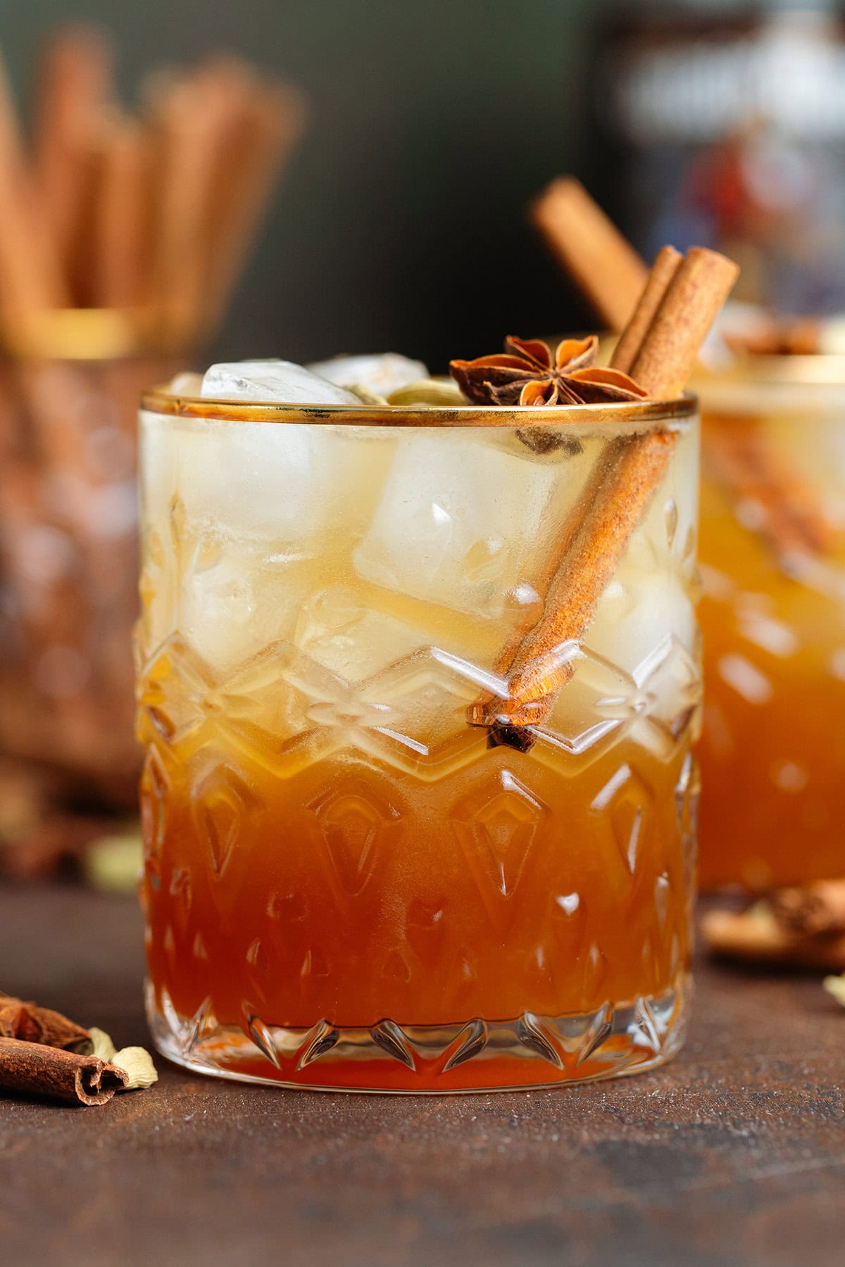 Dark orange cocktail with ice and sparkling water on top for a layered effect in a short glass with a gold rim garnished with a cinnamon stick and star anise.