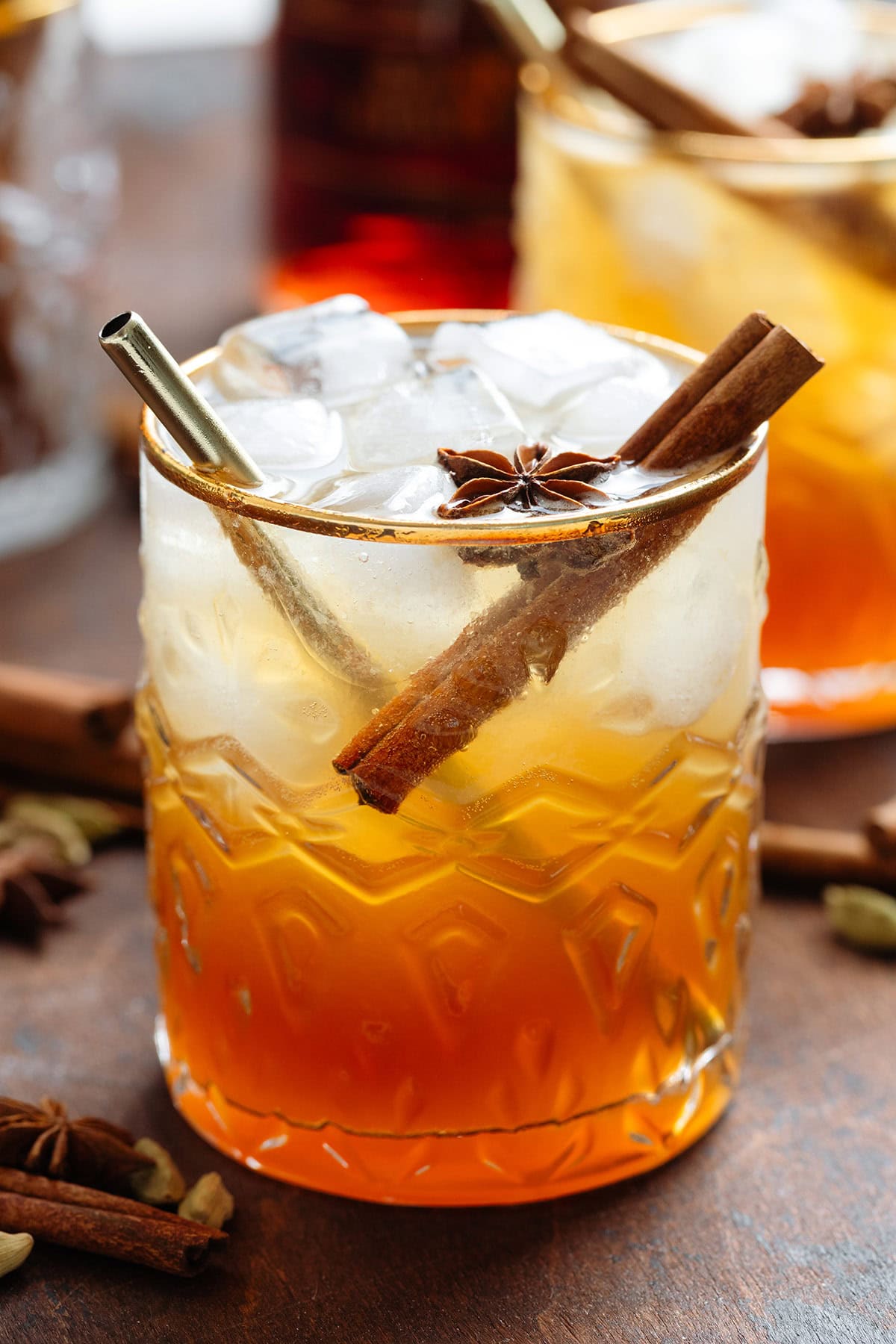 Dark orange cocktail with ice and sparkling water on top for a layered effect in a short glass with a gold rim garnished with a cinnamon stick and star anise.