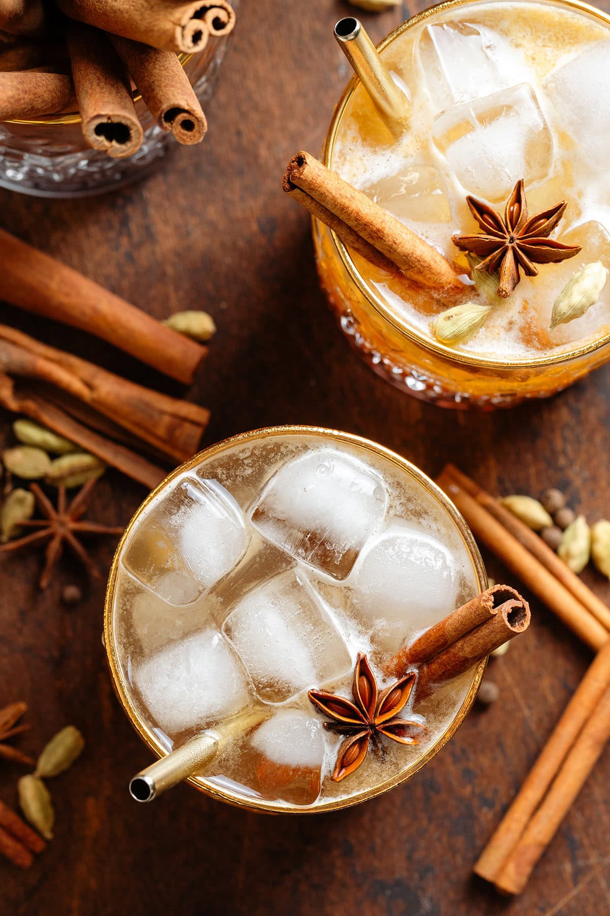 Dark orange cocktail with ice in a glasses with a gold rim garnished with a cinnamon sticks, star anise, and gold straws.