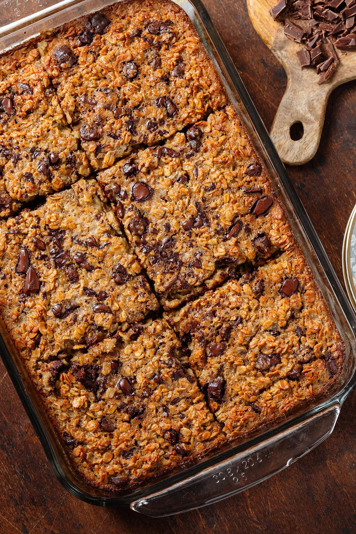 Golden chocolate chip baked oatmeal in a large glass baking dish sliced into 6 squares with more chopped chocolate on a cutting board on the right.