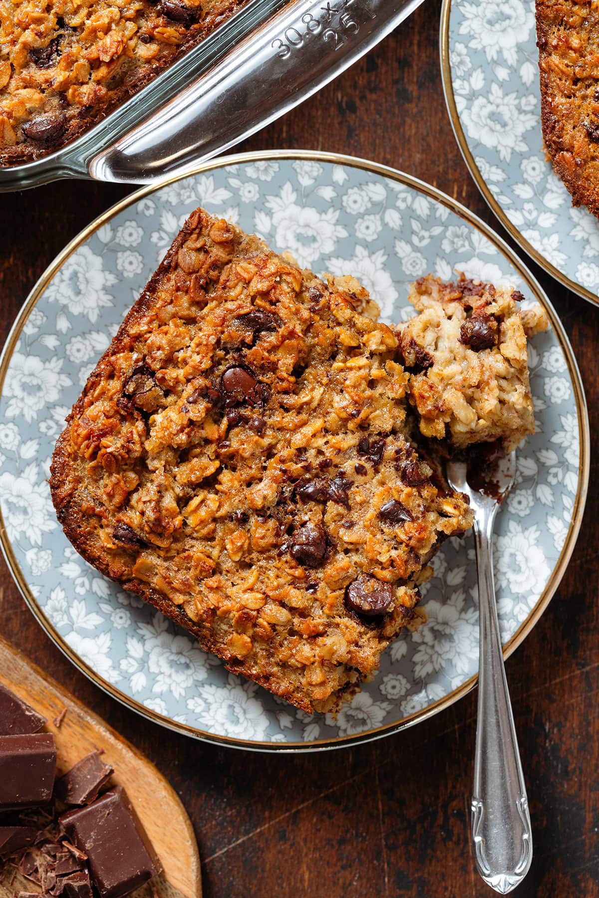 A slice of chocolate chip baked oatmeal on a small blue plate with flowers with a fork on the right holding a bite of the oatmeal.