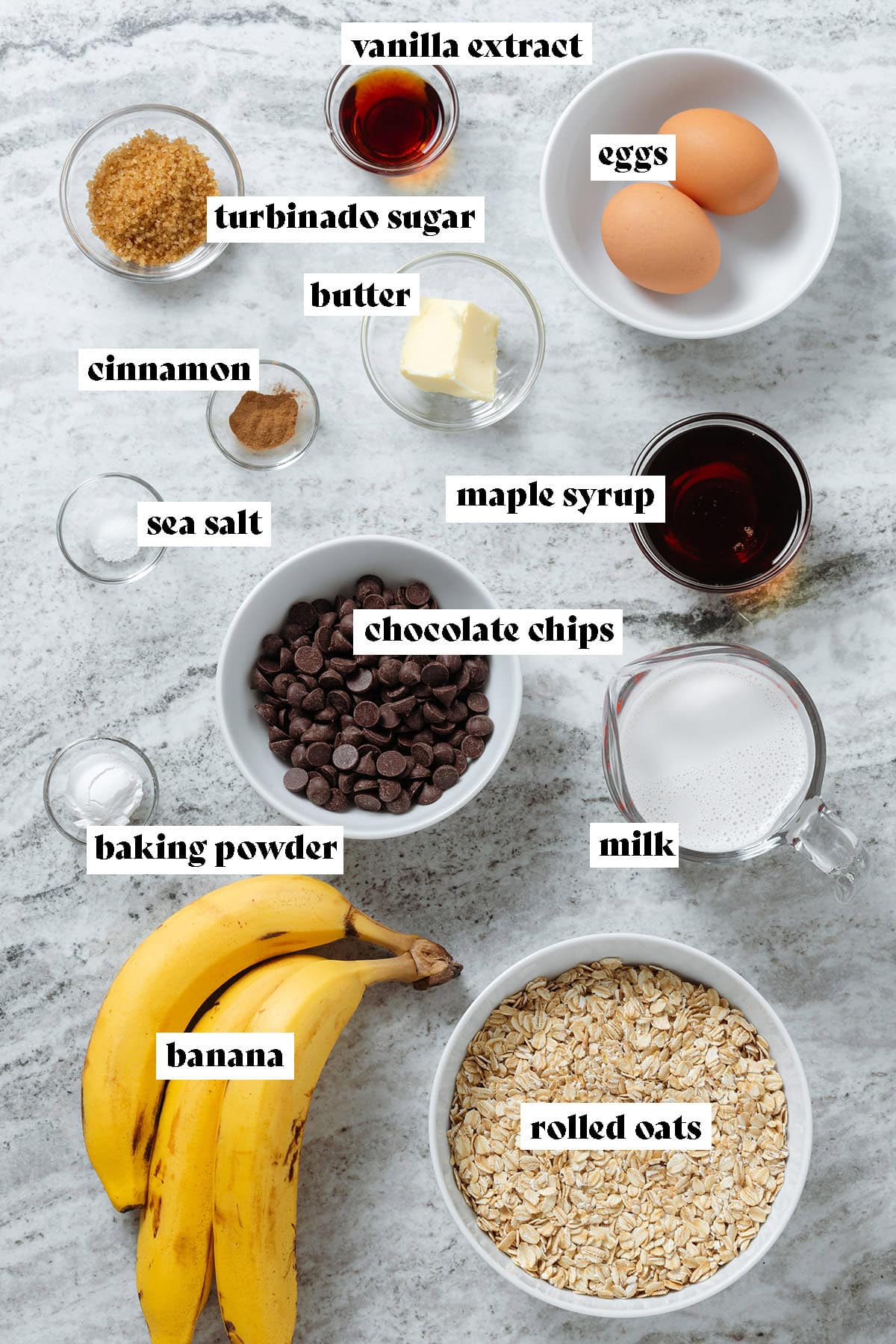 Ingredients like bananas, rolled oats, chocolate chips, milk, maple syrup, and spices in glass bowls of various sizes.