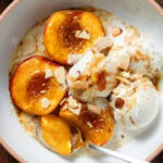 Three baked ad caramelized peach halves in a shallow white bowl topped with three scoops of vanilla ice cream and toasted sliced almonds with a spoon cutting a piece of one peach.