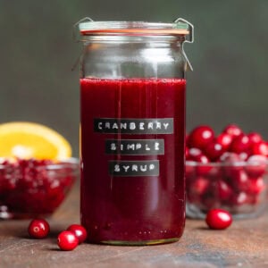 Bright red cranberry syrup in a tall glass jar with more cranberries behind the glass in a bowl.