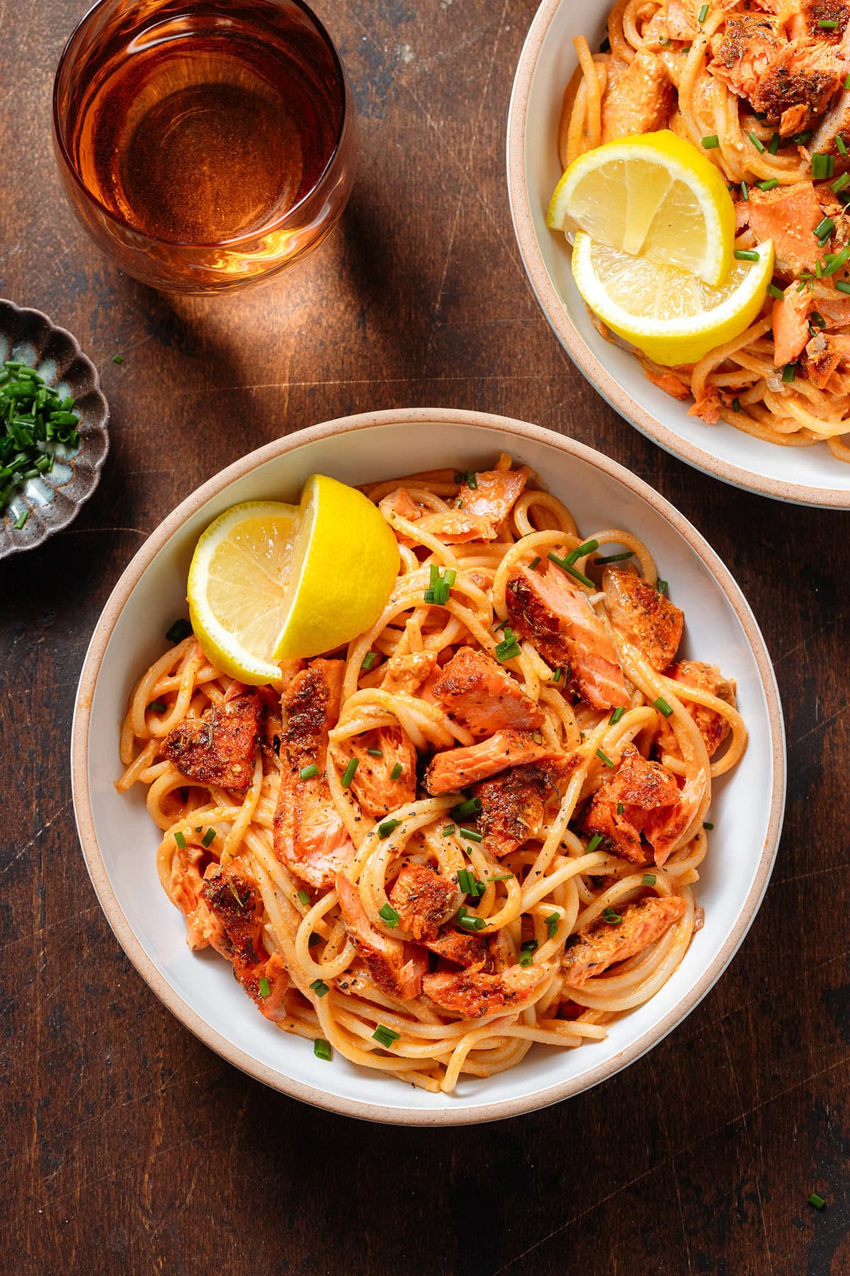 Spaghetti with creamy red sauce and chunks of roasted salmon in white bowls sprinkled with fresh herbs and with a lemon wedges on the side of the bowls.