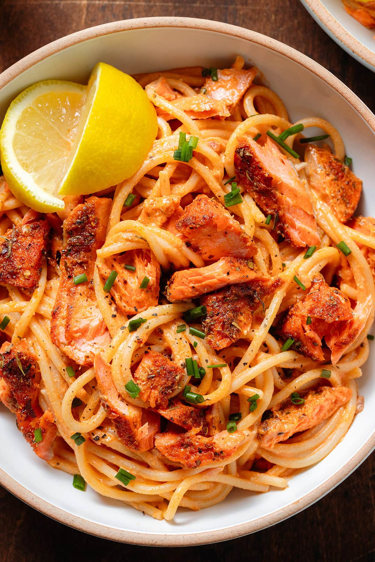 Spaghetti with creamy red sauce and chunks of roasted salmon in a white bowl sprinkled with fresh herbs and with a lemon wedges on the side.