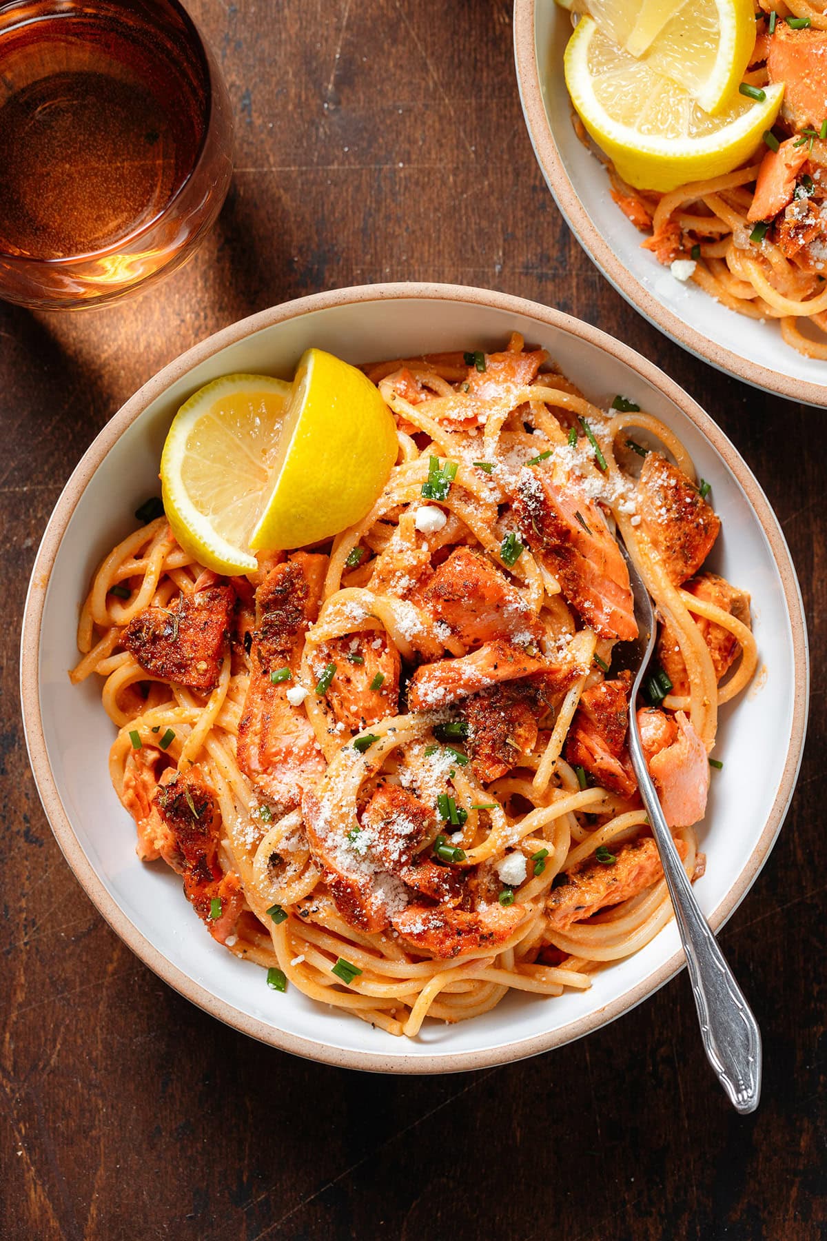 Spaghetti with creamy red sauce and chunks of roasted salmon in white bowls sprinkled with parmesan, fresh herbs, and with a lemon wedges on the side.