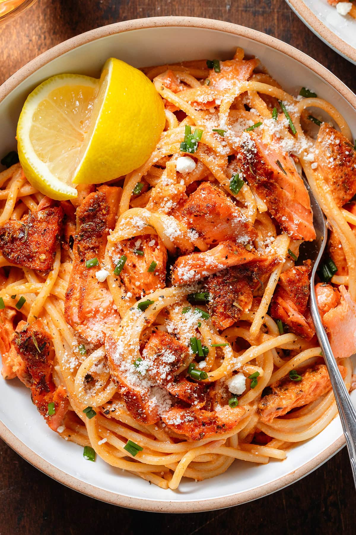 Spaghetti with creamy red sauce and chunks of roasted salmon in white bowls sprinkled with parmesan, fresh herbs, and with a lemon wedges on the side.