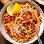 Spaghetti with creamy red sauce and chunks of roasted salmon in a white bowl sprinkled with parmesan, fresh herbs, and with a lemon wedges on the side.