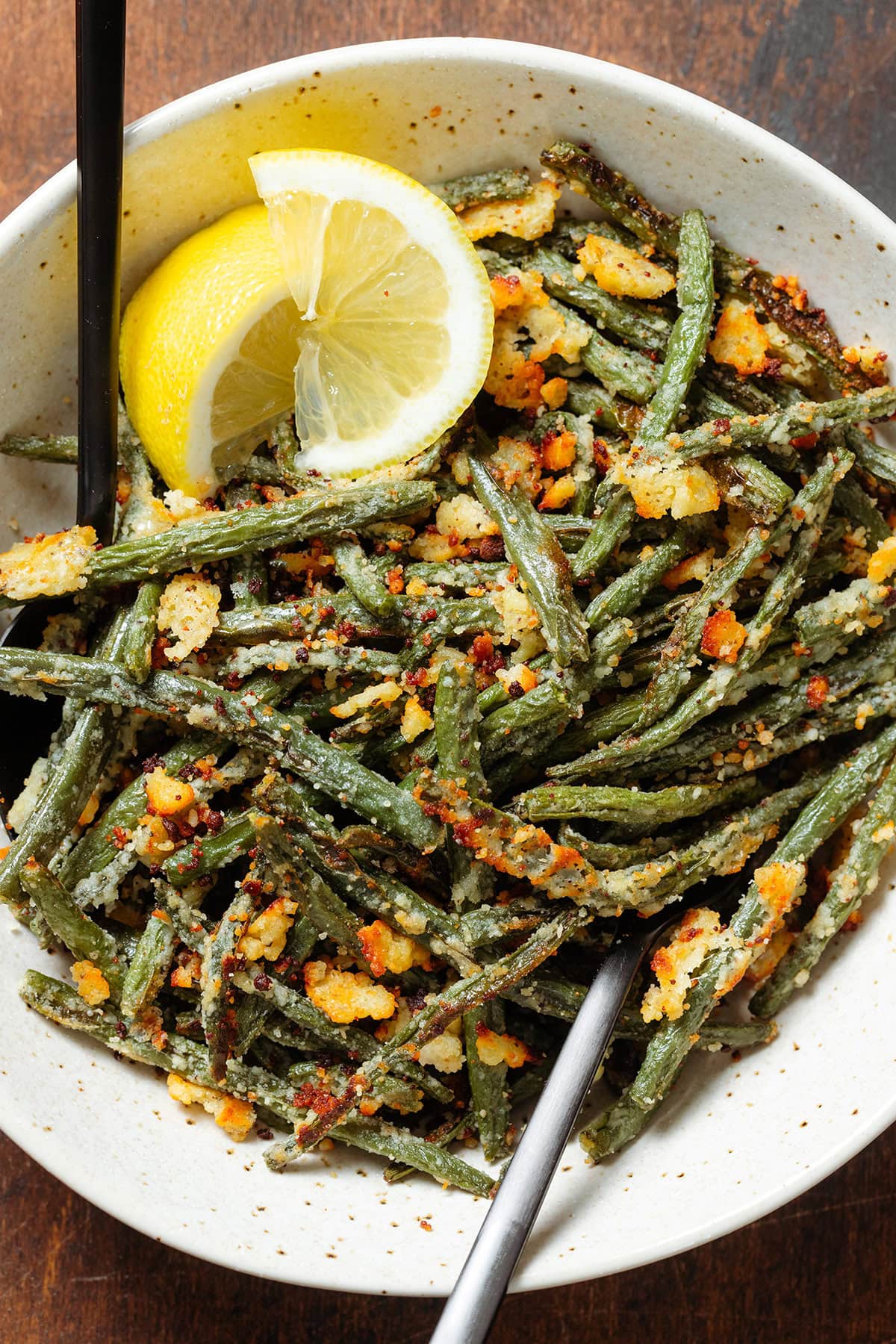 Roasted green beans with crispy parmesan in a beige bowl with two lemon wedges on the left side and serving spoons.