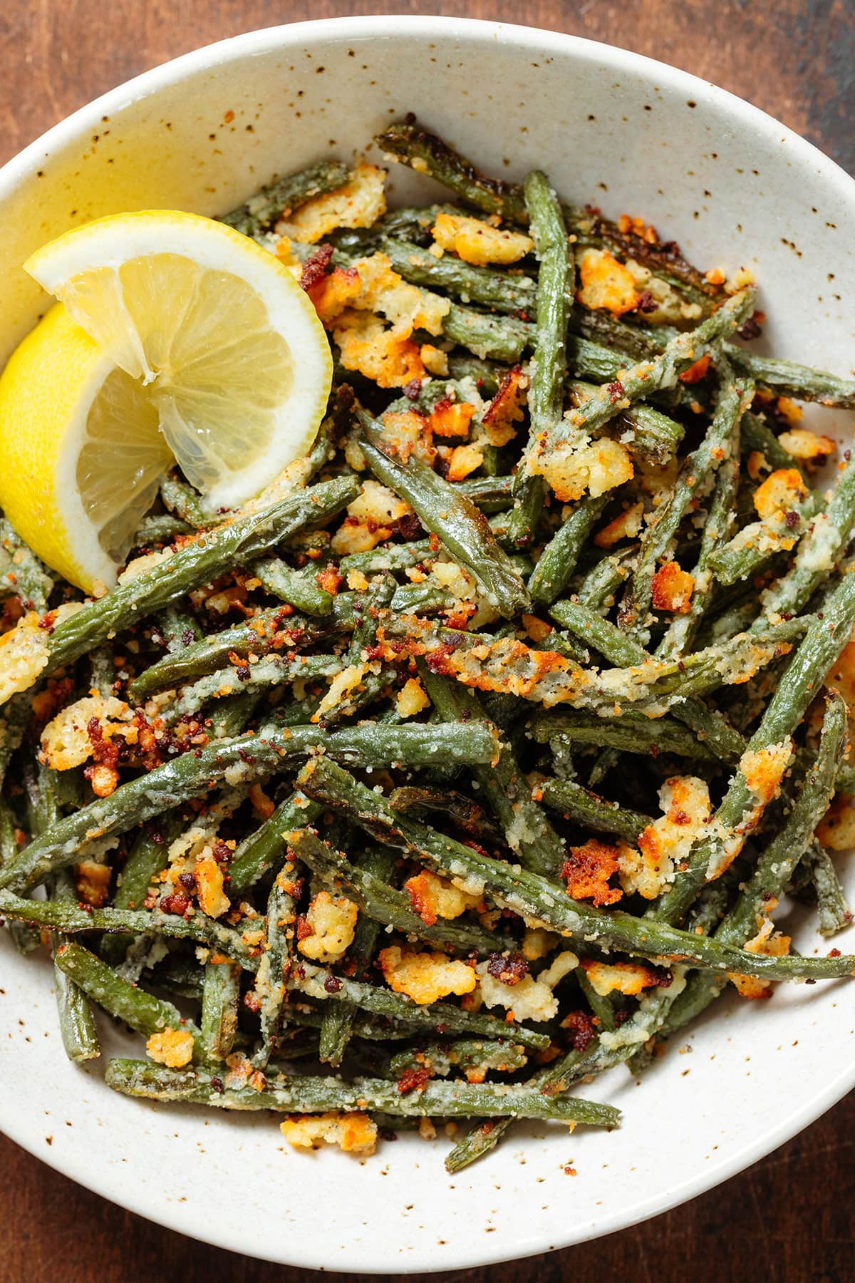 Roasted green beans with crispy parmesan in a beige bowl with two lemon wedges on the left side.