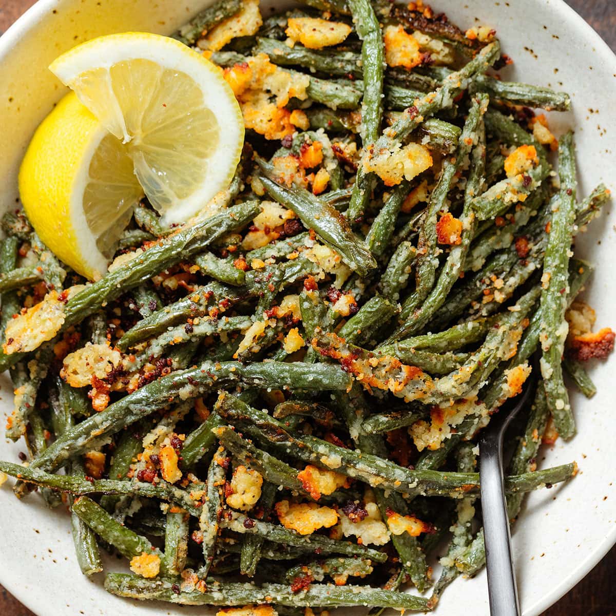 Roasted green beans with crispy parmesan in a beige bowl with two lemon wedges on the left side.