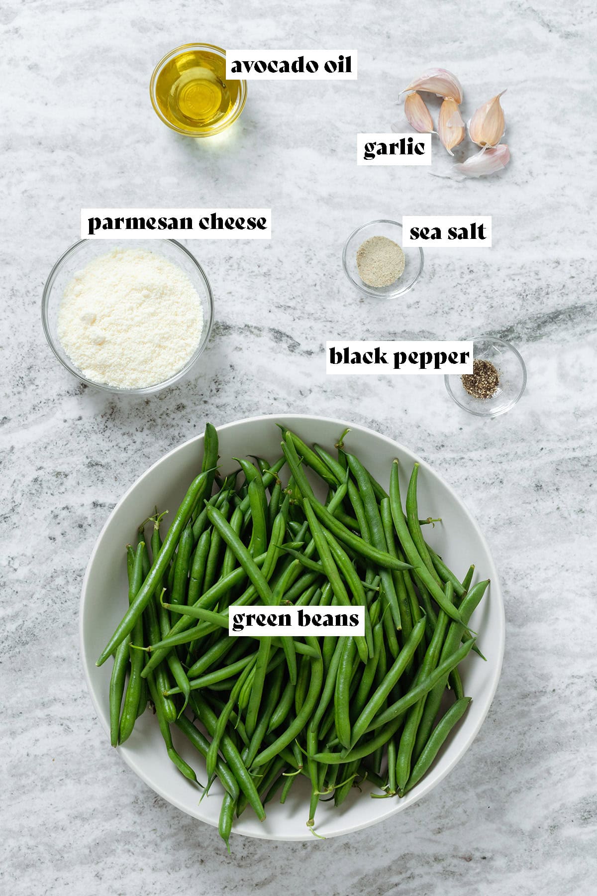 Fresh green beans in a large bowl, garlic cloves, parmesan cheese, spices and oil in small glass bowls.