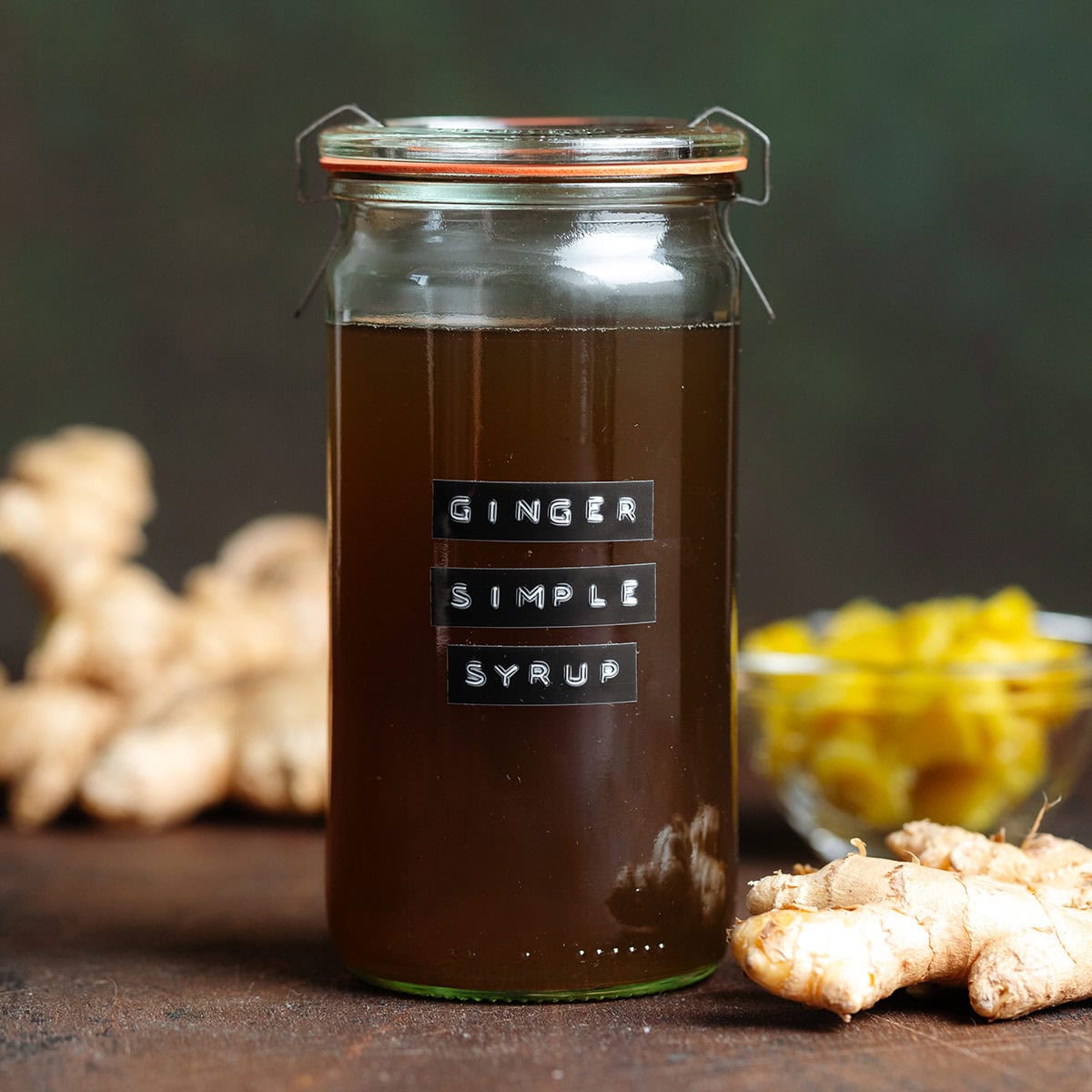 Brown ginger syrup in a tall glass jar with a lid and fresh ginger around the jar on a dark brown background.