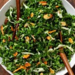 Kale salad with shredded cabbage, carrots, and toasted almonds in a large white bowls with serving spoons.