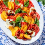 Sliced tomatoes, peaches, mozzarella, and fresh basil layered on a large white serving plate drizzled with balsamic glaze and olive oil.