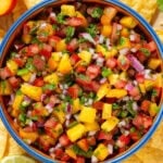 Bright colorful fresh salsa with peaches and tomatoes in a medium bowl with a blue rim with tortilla chips all around.