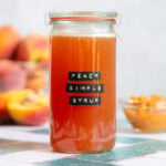 Bright orange peach syrup in a tall glass jar with an embossed label that says peach simple syrup with more peaches behind the glass.