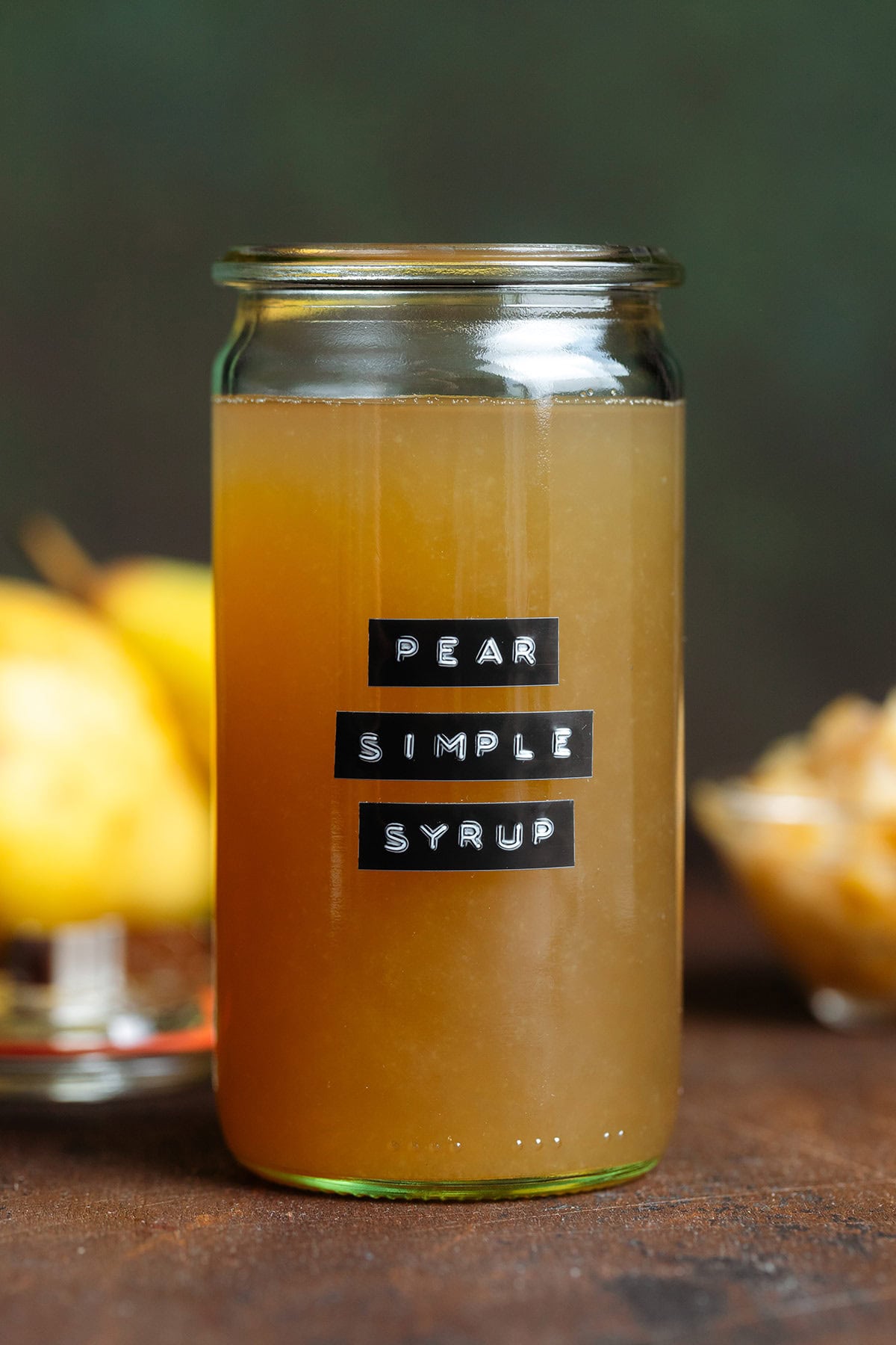 Dark yellow syrup in a glass jar without a lid and with a black embossed label that says pear simple syrup on a dark brown background.