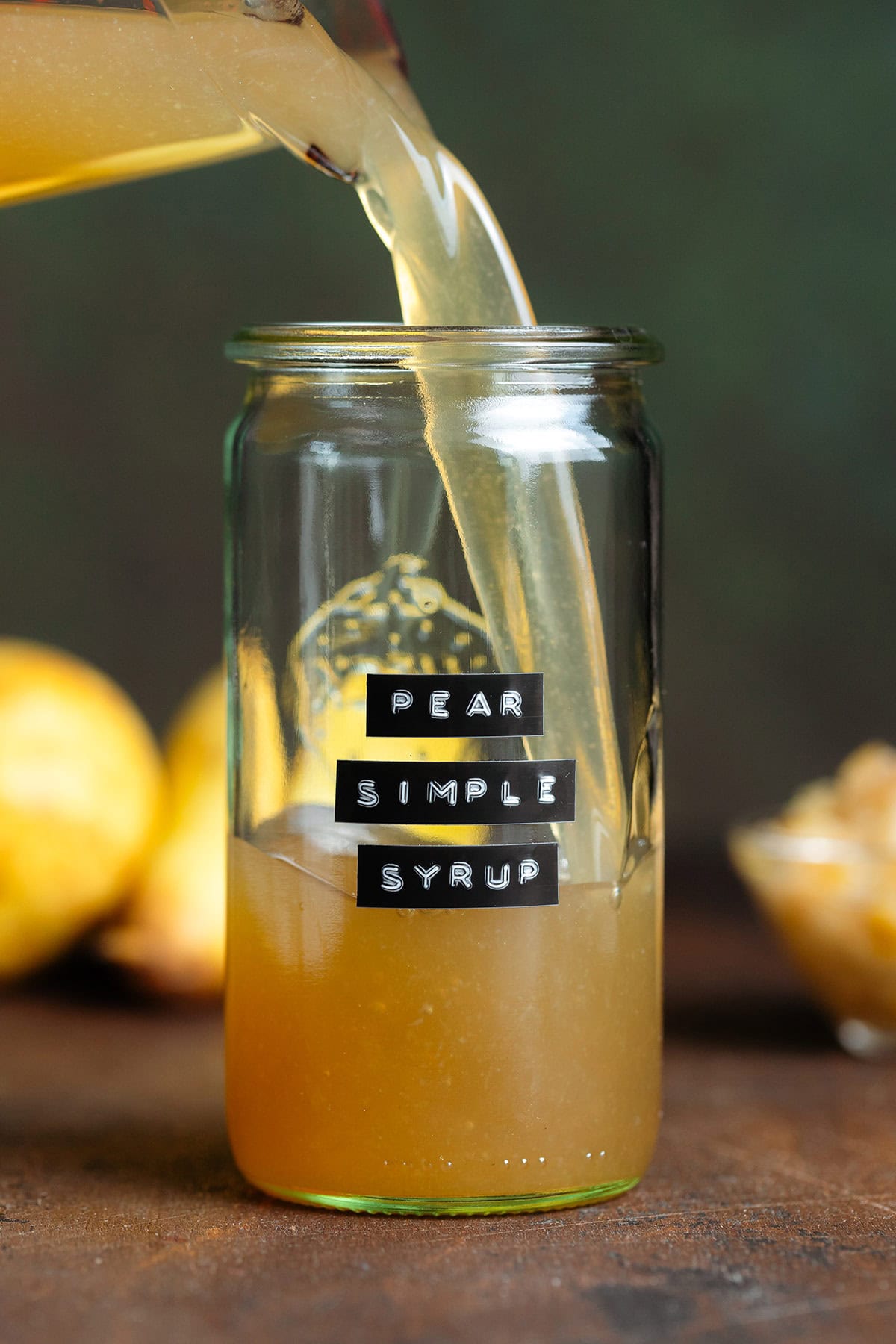 Dark yellow syrup being poured into a tall glass jar on a dark brown background with pears behind the jar.