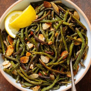 Crispy roasted green beans with toasted sliced almonds in a white bowl with two lemon wedges on the side and a spoon on the right.