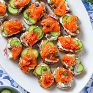 Crostini with cream cheese, smoked salmon, cucumber slices, and red onion on a large white serving plate.