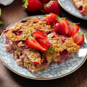 A square of baked oatmeal on a small blue plate with flower motive with more chopped fresh strawberries on top.