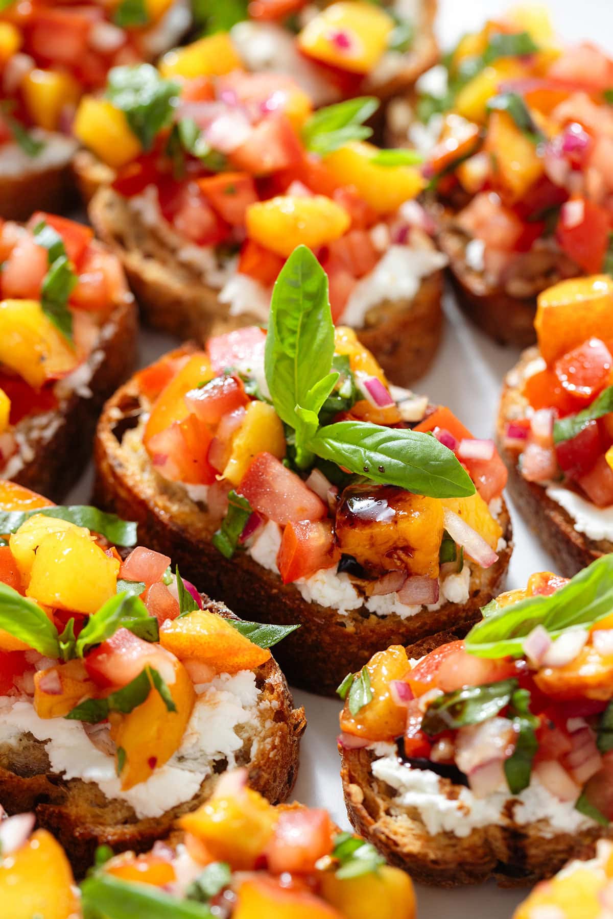 Peach and tomato bruschetta with creamy goat cheese spread and fresh basil drizzled with balsamic glaze on a large white serving plate.