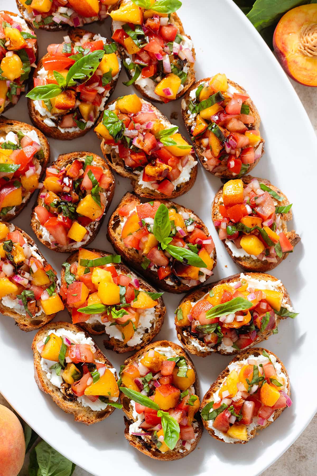 Peach and tomato bruschetta with creamy goat cheese spread drizzled with balsamic glaze on a large white serving plate.