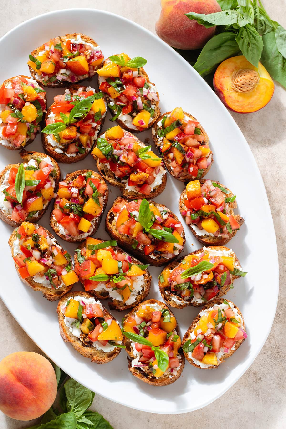 Peach and tomato bruschetta with creamy goat cheese spread and fresh basil drizzled with balsamic glaze on a large white serving plate.