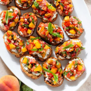 Peach and tomato bruschetta with creamy goat cheese spread drizzled with balsamic glaze on a large white serving plate.