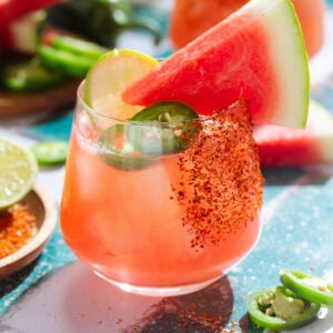 Bright red watermelon margarita in a short glass garnished with fresh watermelon wedge, jalapeno slices, a lime slice, and tajin on one side of the glass.