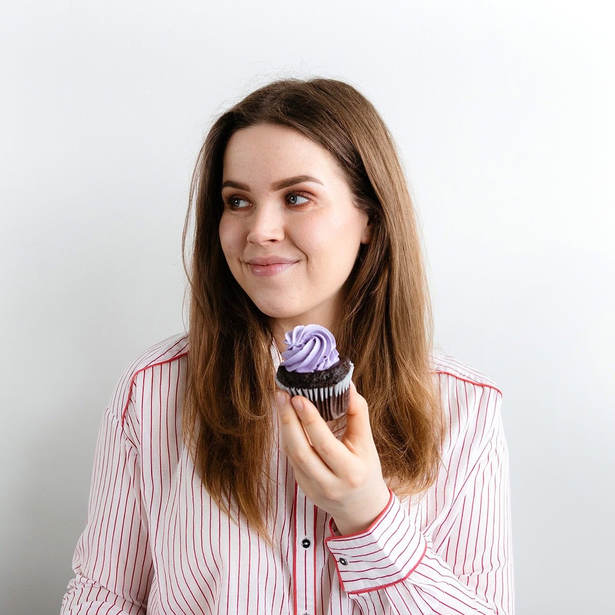 A photo of a young woman in a white and red striped shirt holding a chocolate cupcake with pruple frosting on a white backround.