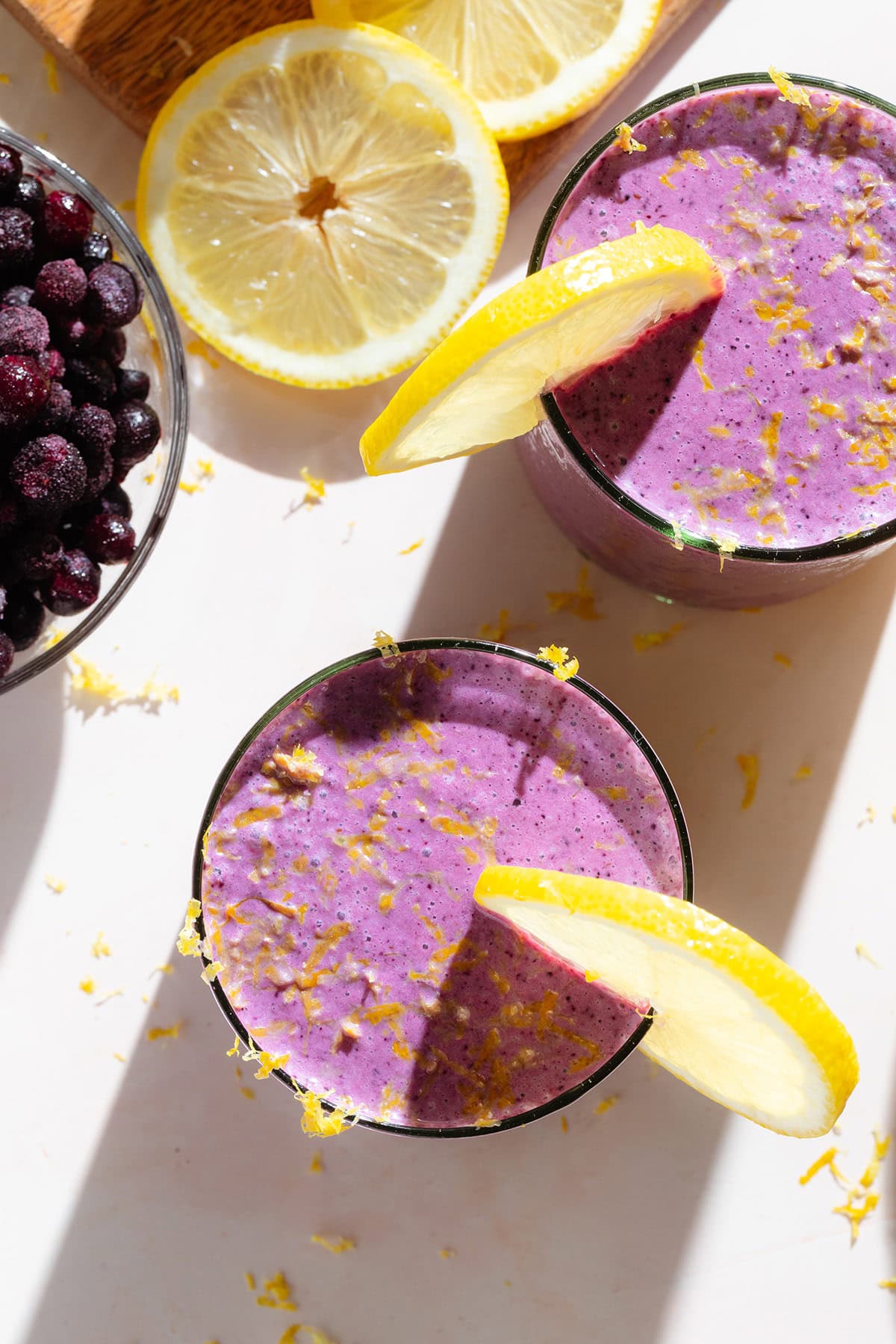 Bright purple smoothie in two glasses garnished with slices of lemon and fresh lemon zest.