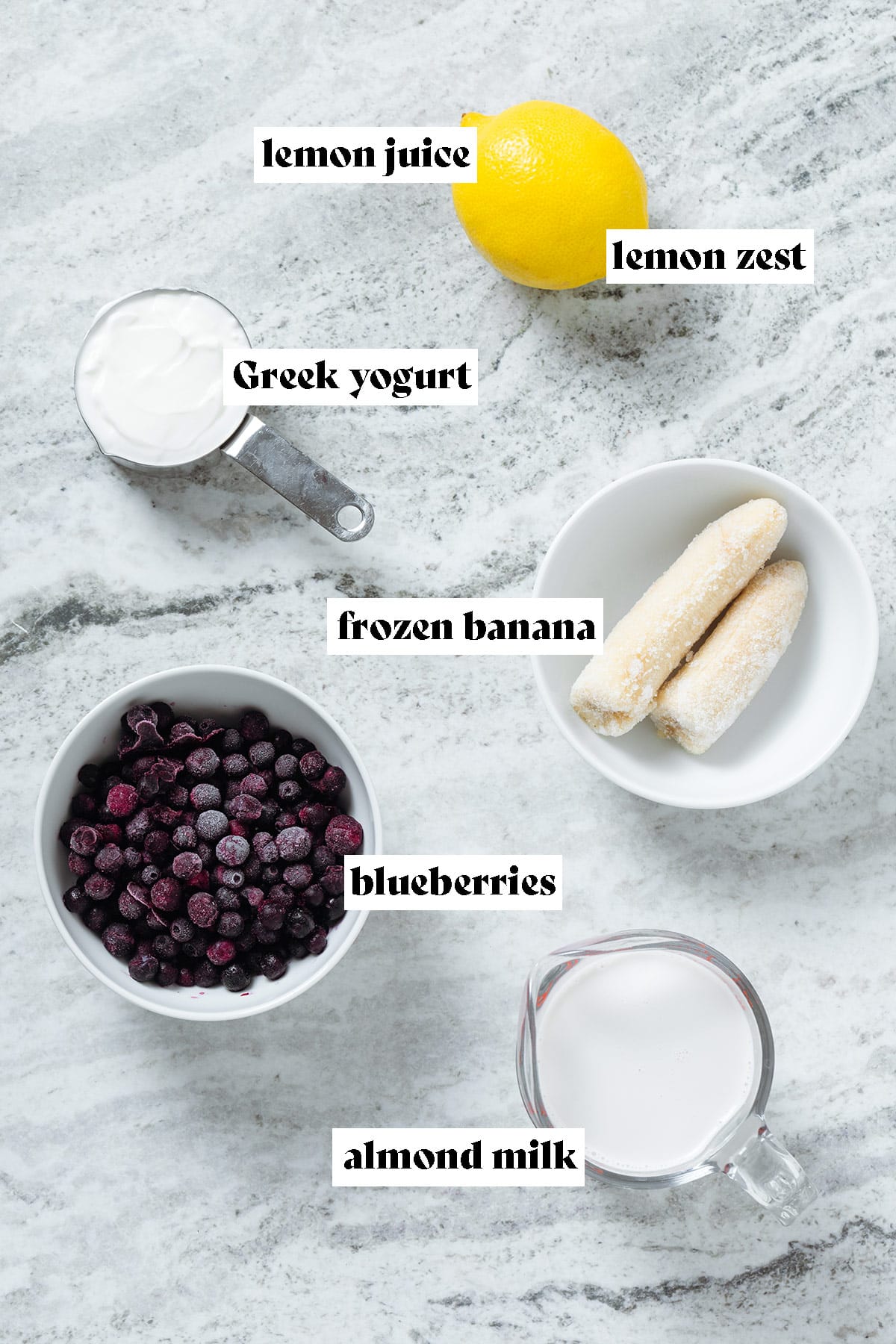 Frozen blueberries, banana, a whole lemon, almon milk, and Greek yogurt laid out in small bowls.