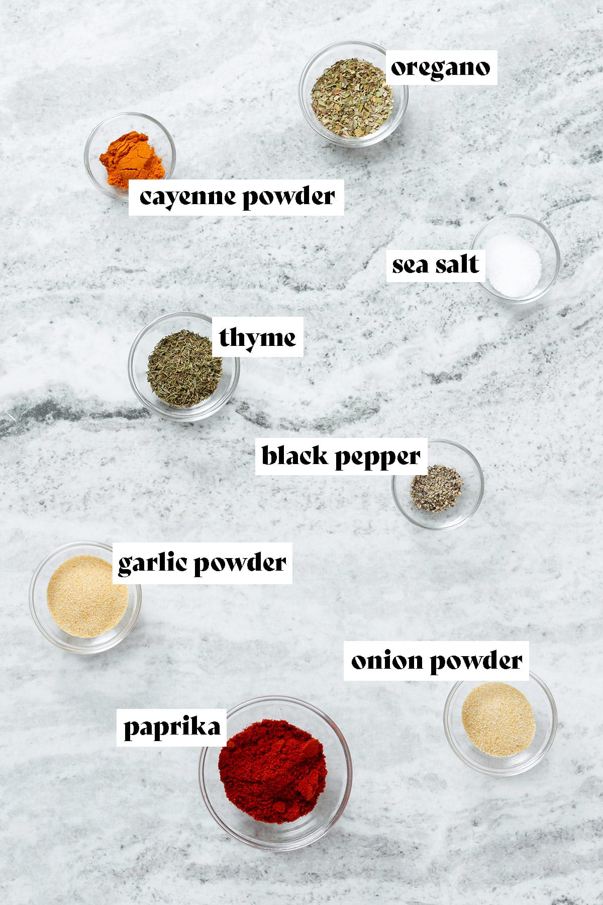 Spices like paprika, garlic powder, oregano, thyme, and sea salt in small glass bowls with text overlay.
