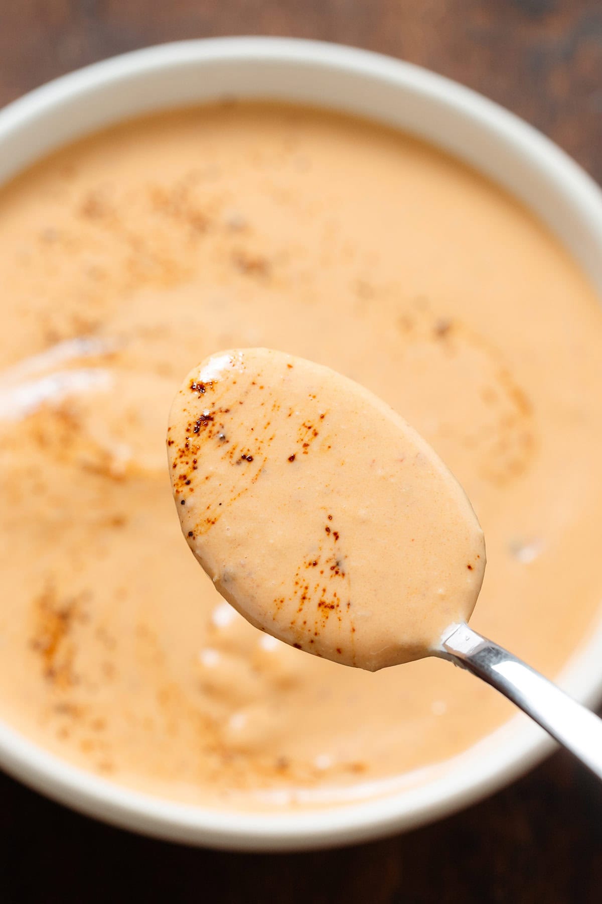 A spoon holding creamy chipotle sauce over a white bowl with the sauce.