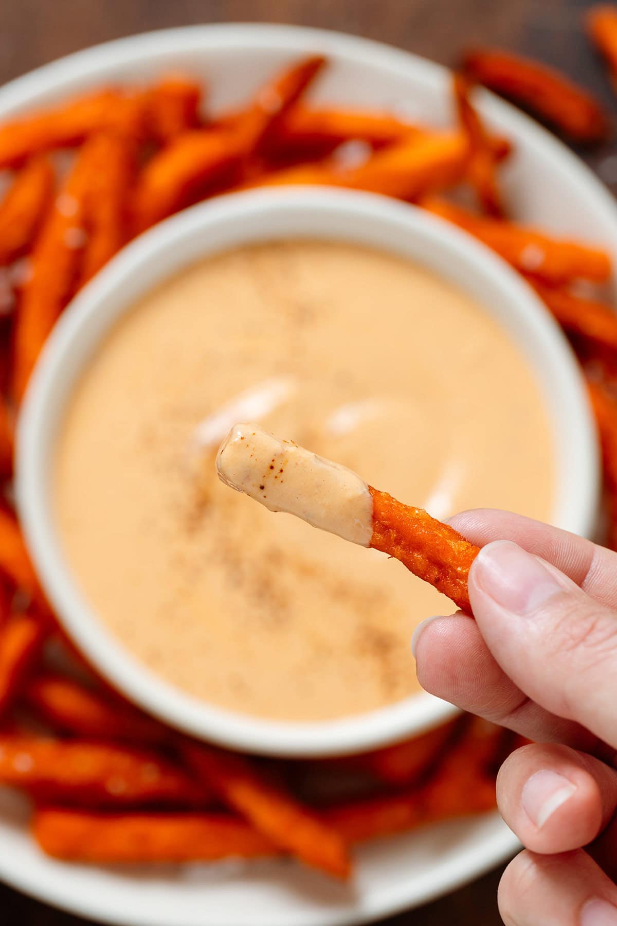 A hand holding a sweet potato fry dipped in creamy orange sauce over a bowl of the sauce surrounded by more fries.