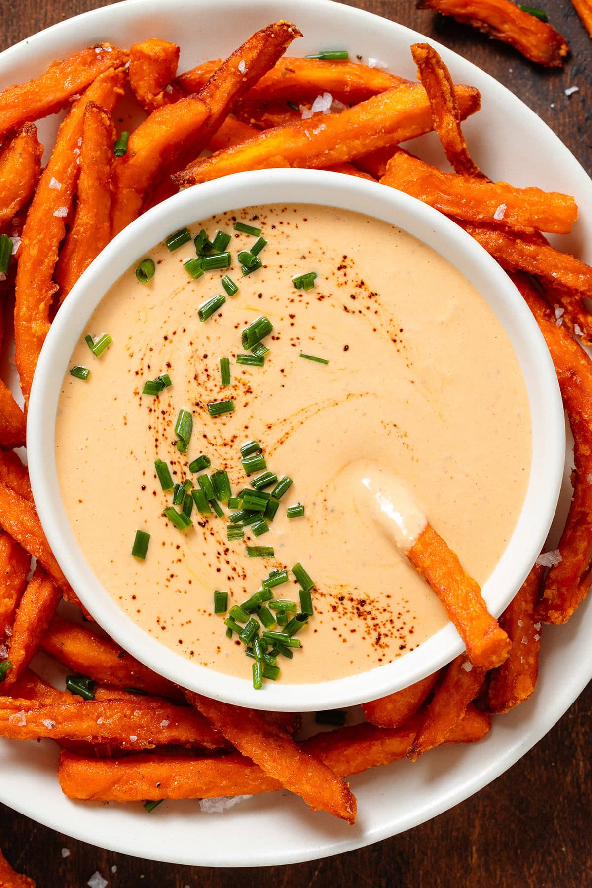 Creamy chipotle sauce in a small white bowl on a plate of sweet potato fries garnished with black pepper and chopped chives.