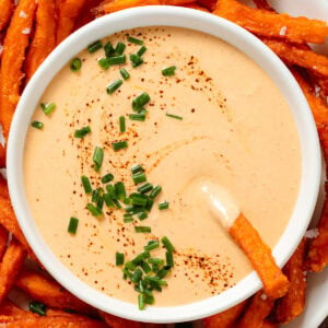 Creamy chipotle sauce in a small white bowl on a plate of sweet potato fries garnished with black pepper and chopped chives.