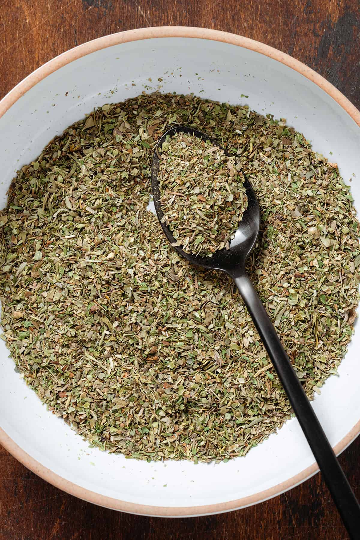 Mixed dry herb seasoning in a low with bowl with a black spoon on the right side of the bowl.