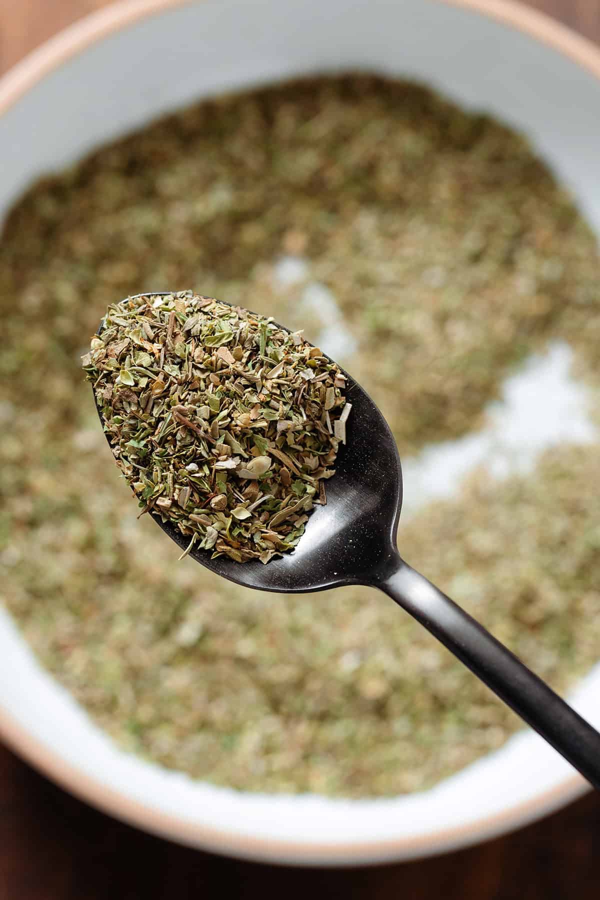 A black spoon holding mixed dried herb seasoning over a white bowl with more of the seasoning.