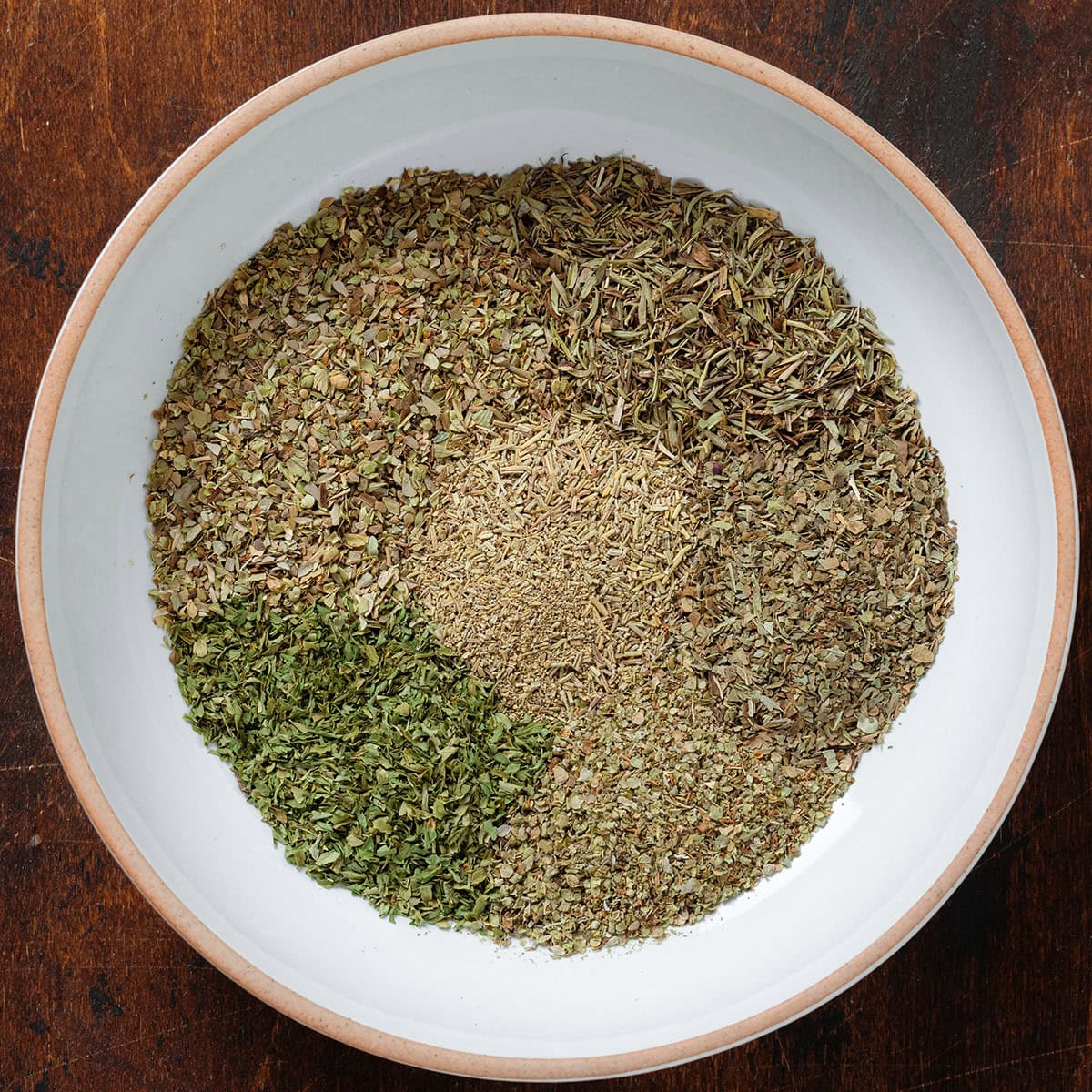 Dried herbs in a low white bowl with an orange rim before being mixed together.