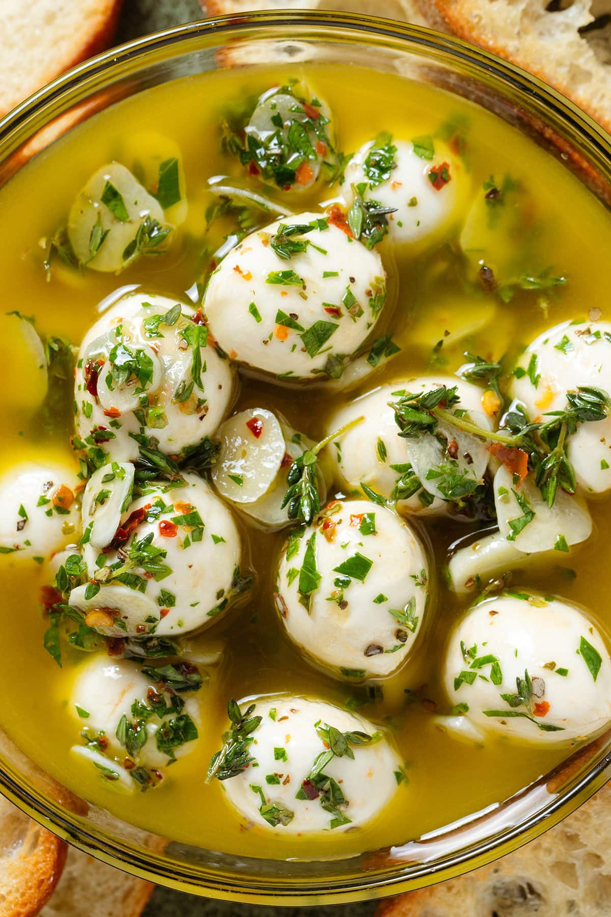 Marinated small mozzarella balls in olive oil and fresh herbs, and sliced garlic in a glass bowl.