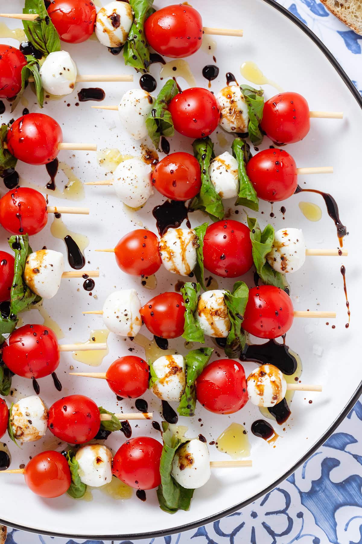 Wooden skewers with cherry tomatoes, bocconcini, and fresh basil drizzled with olive oil and balsamic glaze, and flaky sea salt on a large white platter.